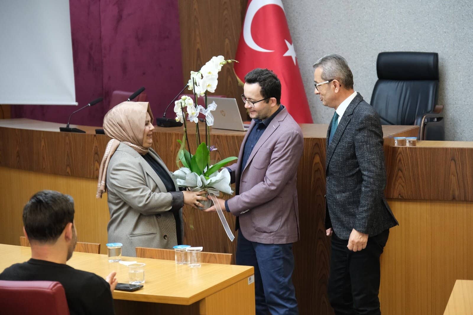 Alternatif Metin: Bir basın toplantısında, bir bayan ve iki erkek arasında çiçek takdimi gerçekleşiyor.