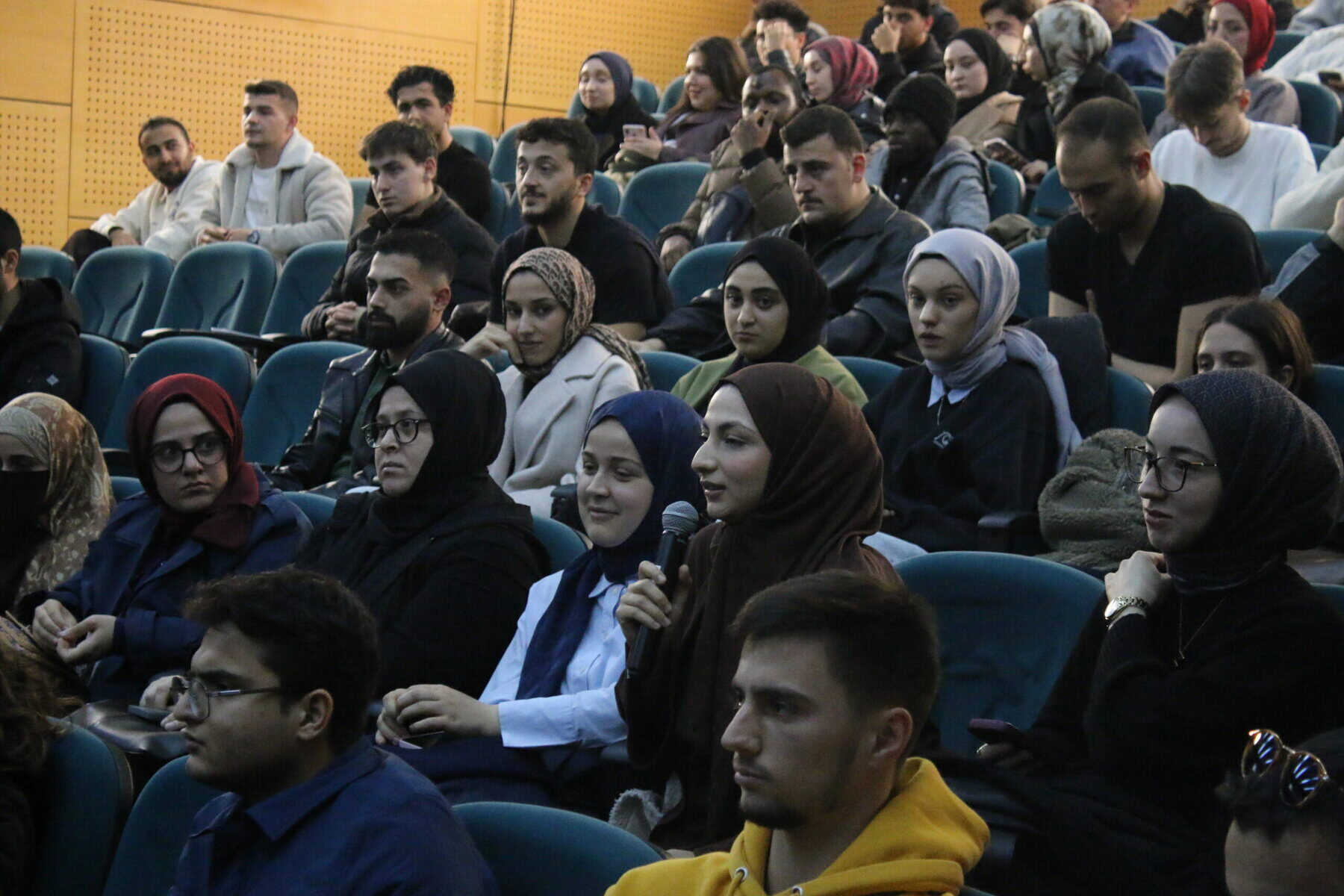 Görselde çeşitli etnik ve yaş grubu insanlardan oluşan kalabalık, konferans veya seminerde yer alıyor.